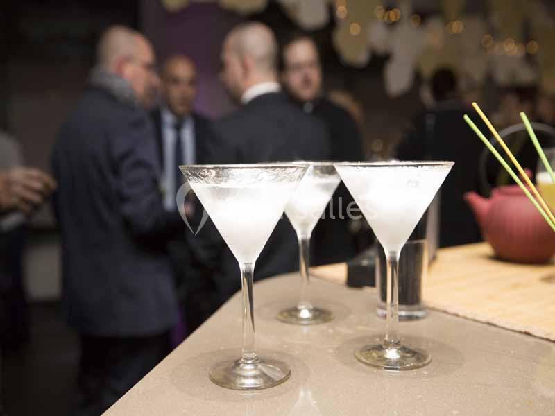 Trois verres à cocktail remplis sur un bar, avec des personnes discutant en arrière-plan flou.