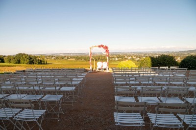 Chaises blanches alignées en extérieur face à une arche décorée de fleurs, avec vue sur un paysage de campagne.