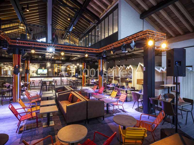 Intérieur d'un bar moderne avec des tables, des chaises colorées et un éclairage chaleureux sous un plafond en bois.