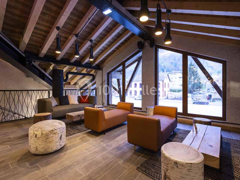 Salon moderne sous combles avec canapés orange, tables basses en bois, poufs clairs et grandes fenêtres donnant sur l…