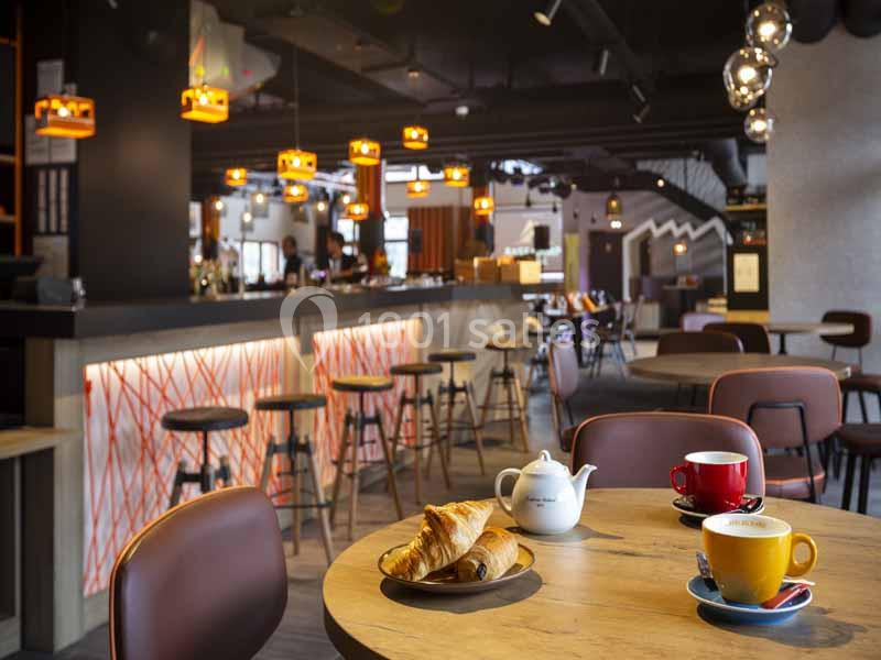 Intérieur d'un café moderne avec un comptoir éclairé, des tabourets et une table garnie de croissants et boissons chaudes.