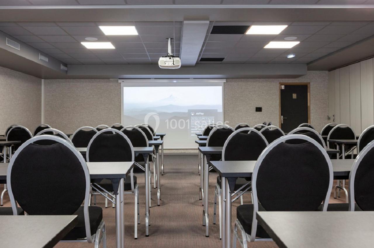 Salle de conférence vide avec des rangées de chaises noires et des tables face à un écran de projection allumé.
