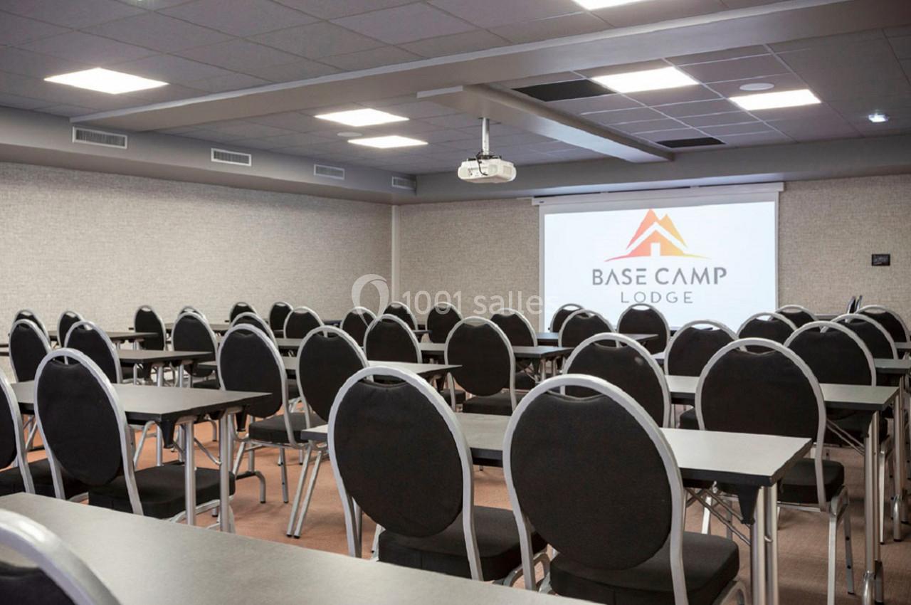 Salle de conférence équipée de tables, chaises et projecteur, avec un écran affichant le logo ’Base Camp Lodge’.