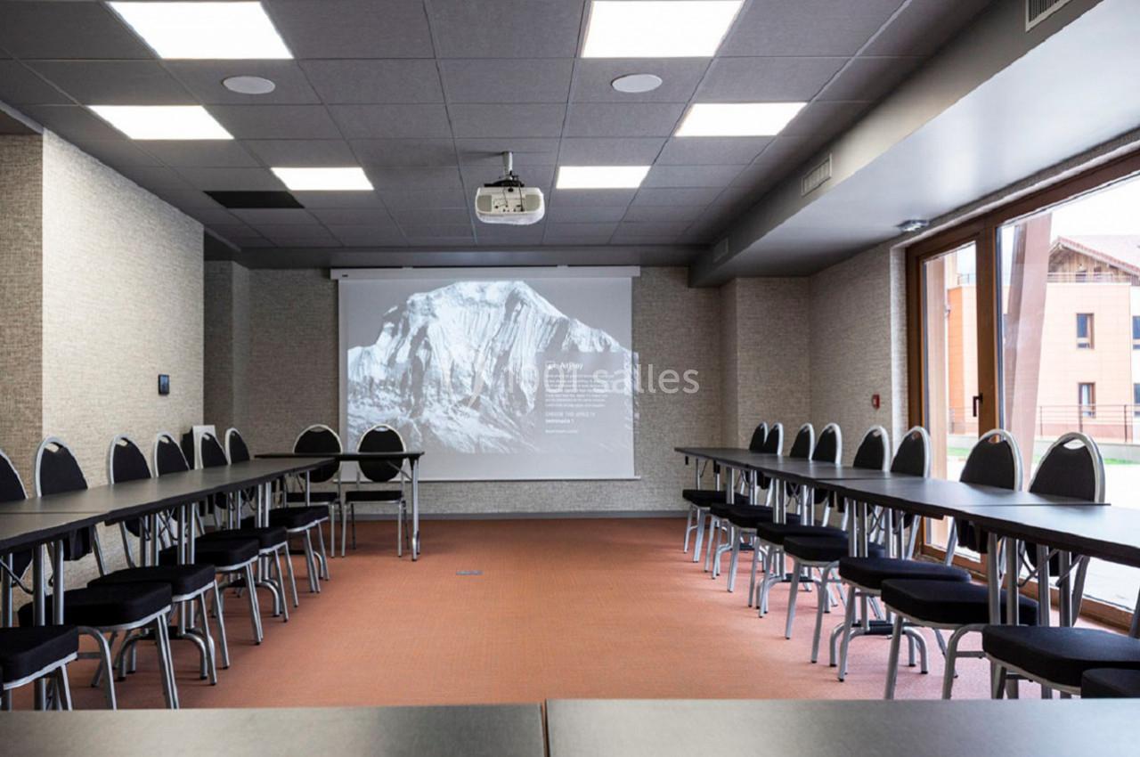 Salle de réunion équipée de tables disposées en U, chaises noires, vidéoprojecteur et écran blanc.