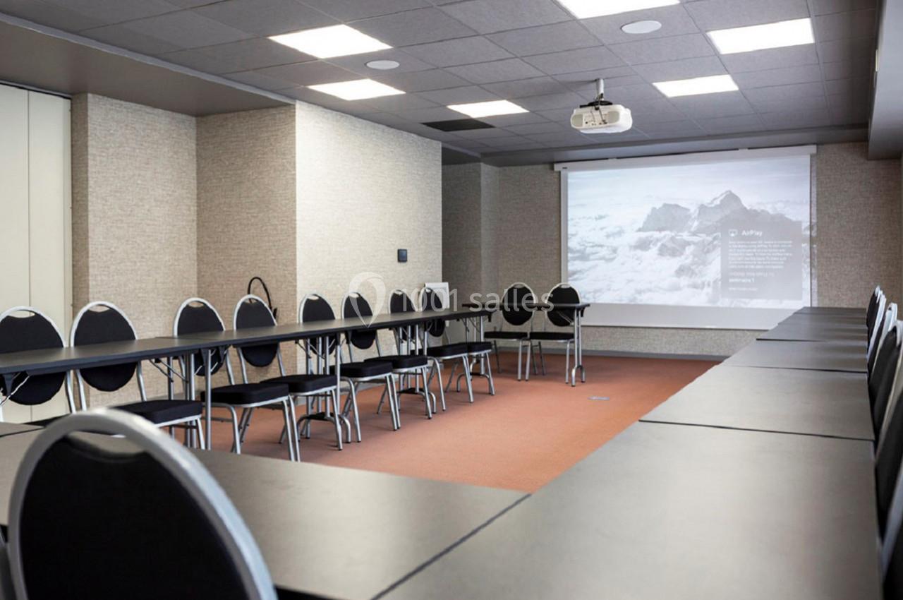 Salle de réunion équipée de tables en U, chaises noires, projecteur et écran affichant une image de montagne.