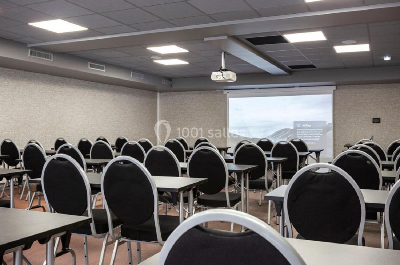 Salle de conférence avec des rangées de chaises noires et tables, écran de projection allumé en arrière-plan.