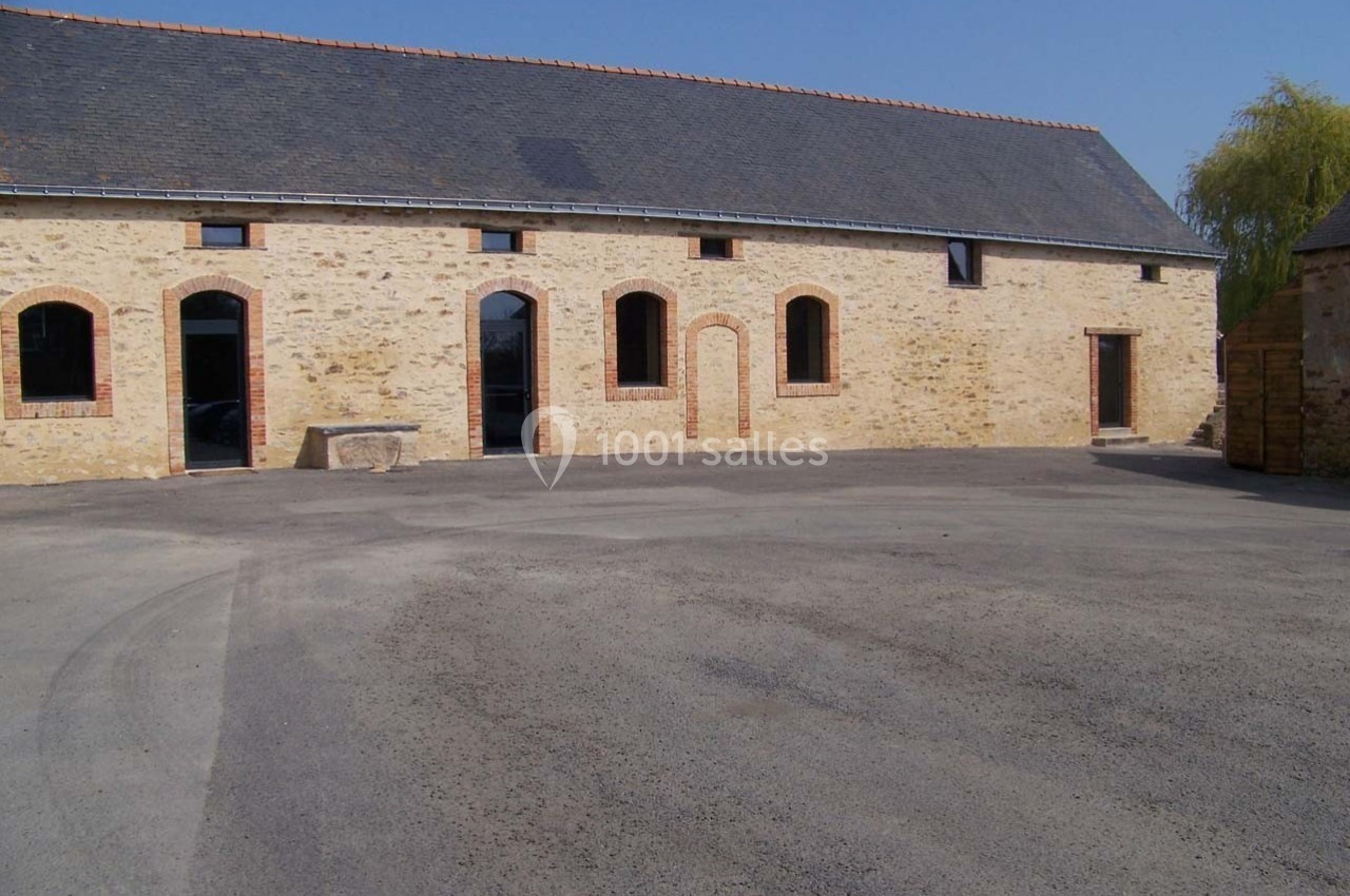 Bâtiment en pierre avec toit en ardoise, grandes ouvertures et cour asphaltée sous un ciel dégagé.