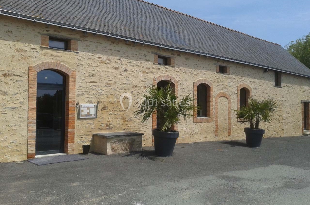 Bâtiment en pierre avec toit en ardoise, grandes ouvertures en arc et deux palmiers en pots devant l'entrée.