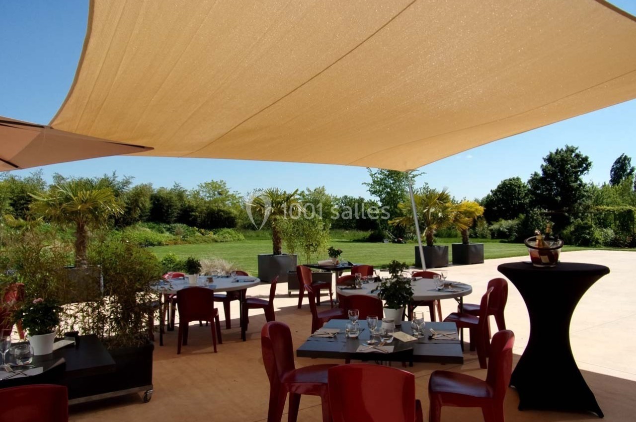 Terrasse extérieure avec tables et chaises rouges sous une grande voile d'ombrage, entourée de verdure.