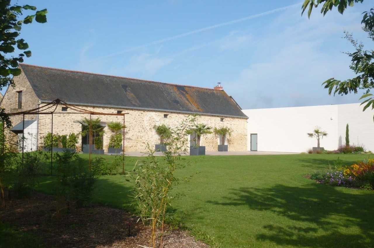 Bâtiment en pierre avec toit en ardoise, entouré d'un jardin verdoyant et de parterres fleuris.