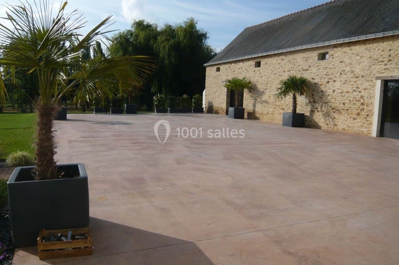Cour spacieuse en béton lisse entourée de palmiers en pots, avec un bâtiment en pierre et un jardin en arrière-plan.