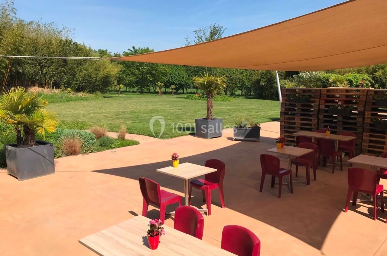 Terrasse avec tables et chaises rouges sous une voile d'ombrage, donnant sur un jardin verdoyant.