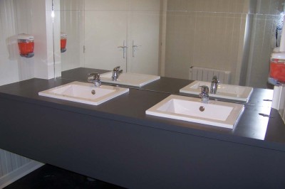 Deux lavabos blancs encastrés dans un plan de travail sombre, avec miroirs et distributeurs de savon muraux rouges.