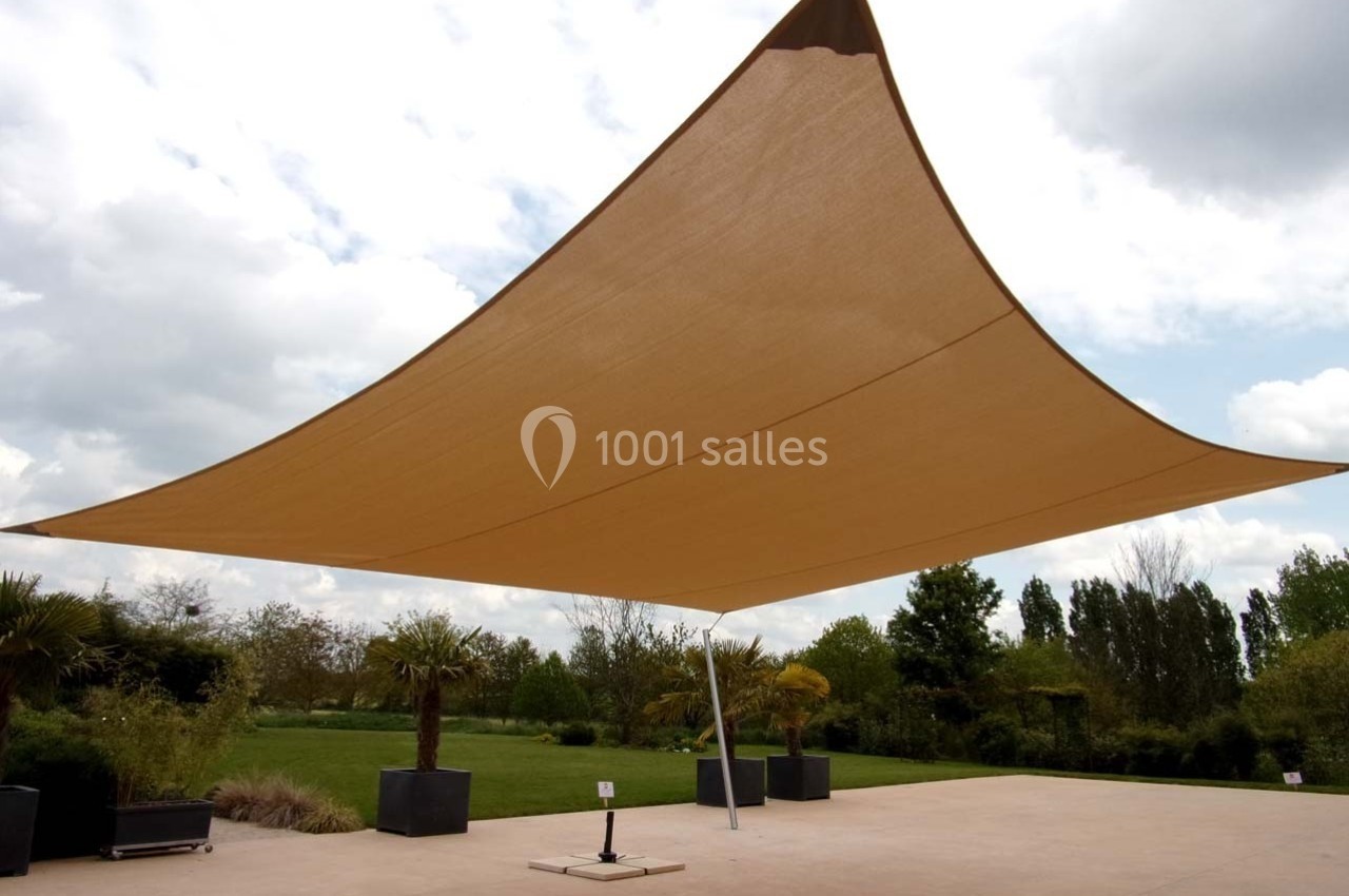Voile d'ombrage beige tendue sur une terrasse extérieure, entourée de végétation et de pots de plantes.