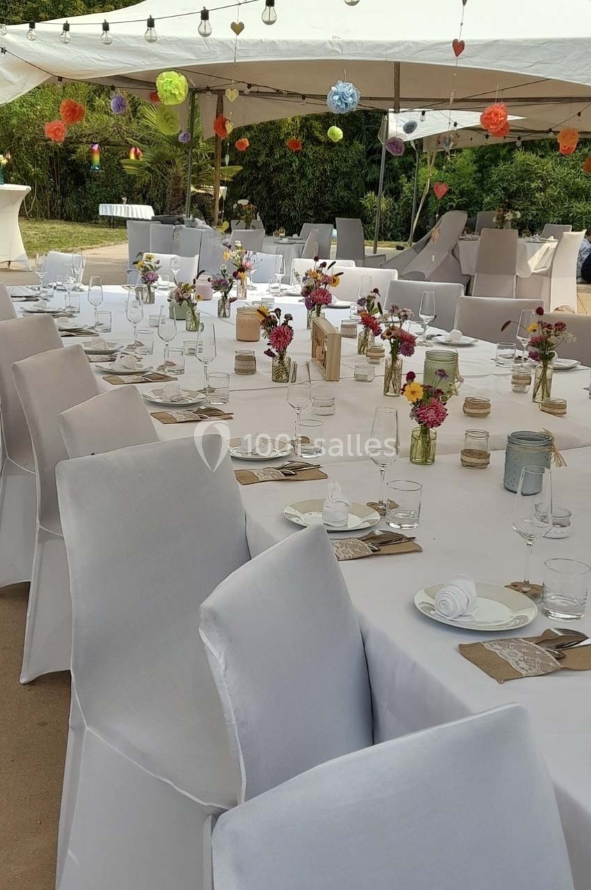 Tables de réception décorées avec nappes blanches, fleurs colorées et lanternes, sous une tente en extérieur.