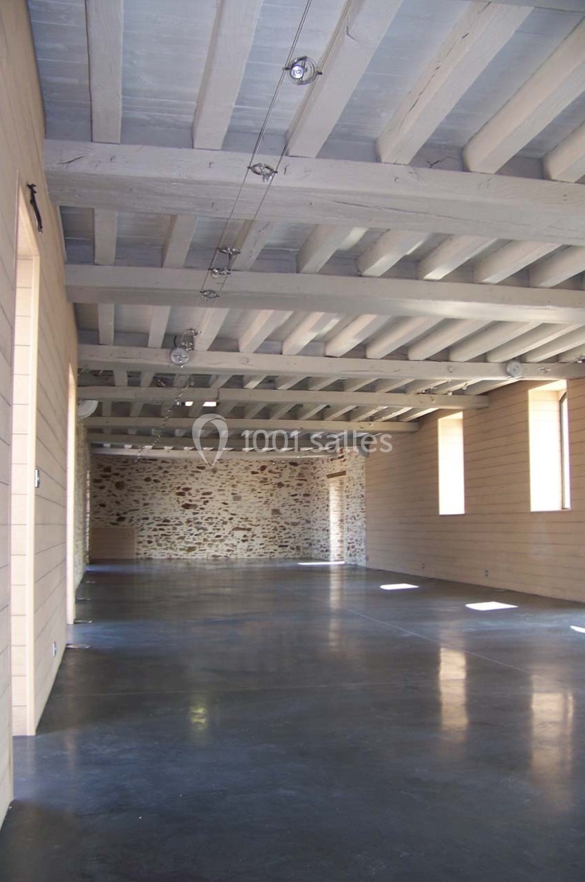 Salle vide avec murs en bois clair, mur en pierre au fond, plafond avec poutres apparentes et sol en béton poli.