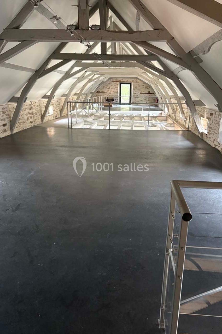 Vue d'un espace intérieur rénové sous combles avec poutres apparentes, sol sombre et mezzanine en verre.