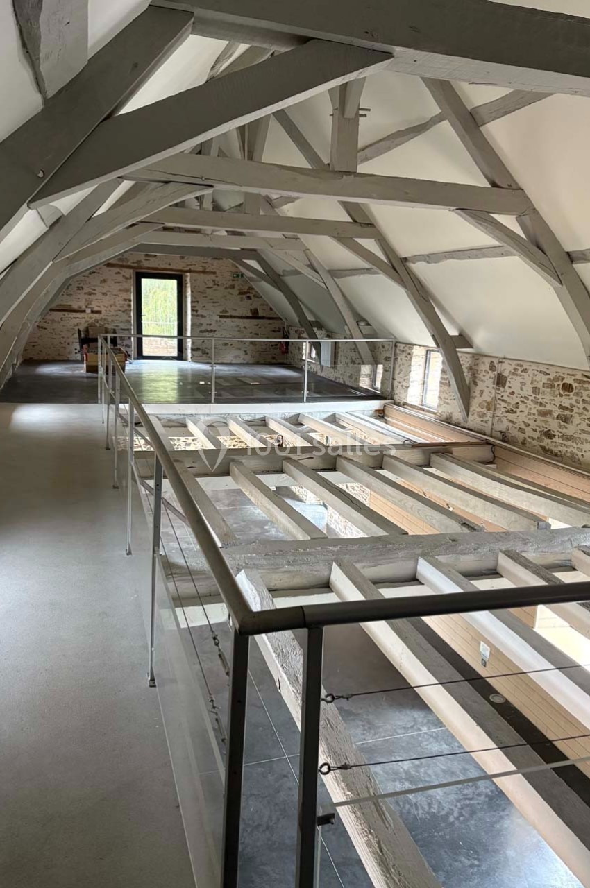 Vue intérieure d'un grenier rénové avec poutres apparentes, murs en pierre et garde-corps en verre.