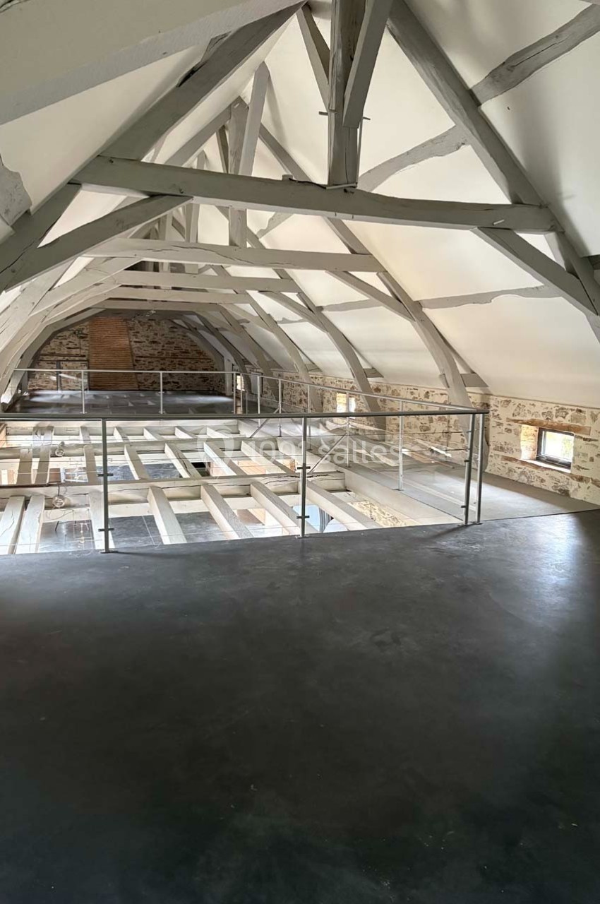 Charpente apparente dans un grenier rénové avec murs en pierre, sol en béton et garde-corps métallique.