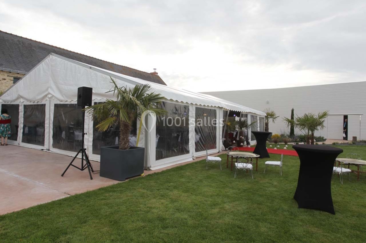 Vue d'une tente blanche aménagée pour un événement, entourée de tables, chaises et palmiers sur une pelouse.