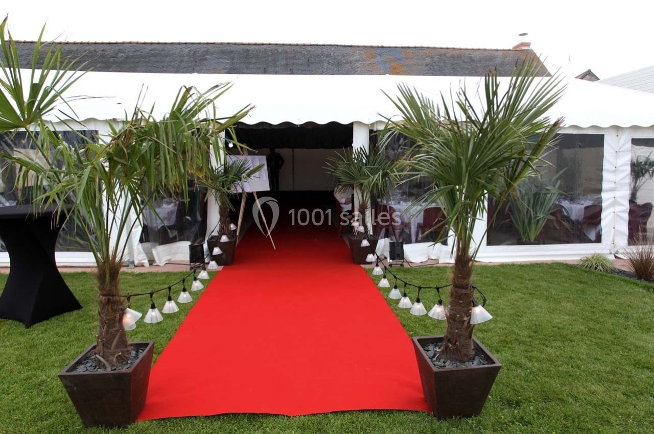 Entrée d'une tente événementielle blanche avec tapis rouge, bordée de palmiers en pots et éclairages au sol.