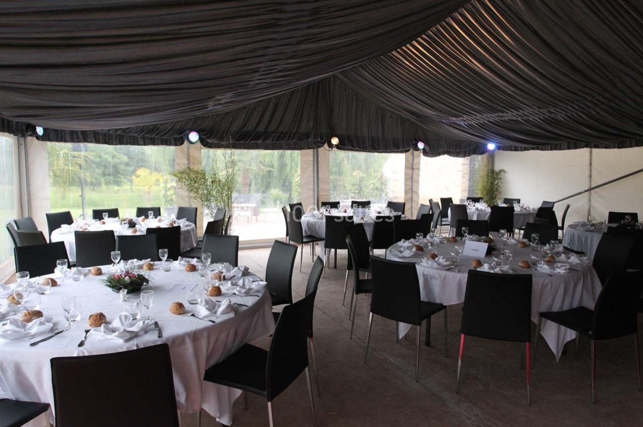 Salle de réception avec tables rondes dressées, nappes blanches, chaises noires et lumière naturelle en arrière-plan.