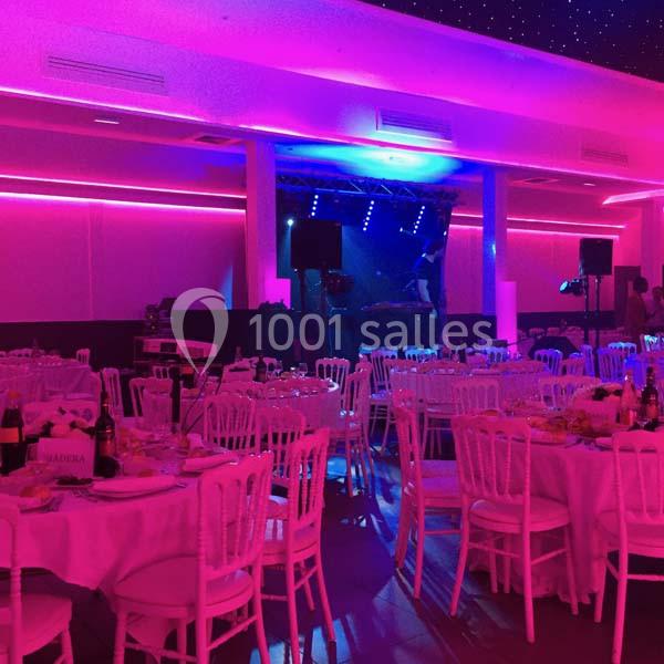 Salle de réception éclairée de lumières roses et violettes, tables dressées avec chaises blanches.