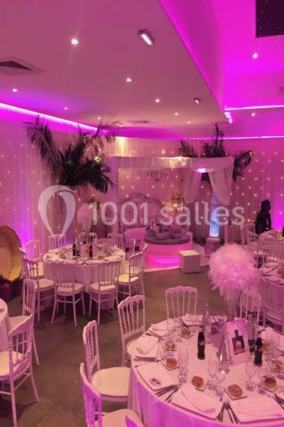 Salle de réception décorée avec des tables rondes, des chaises blanches, des lumières roses et des éléments décoratifs…