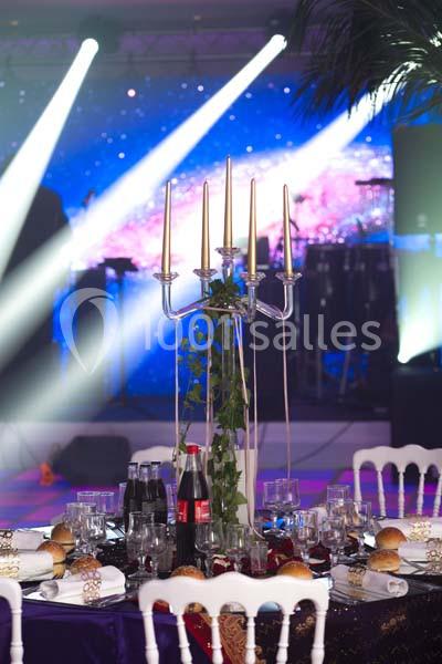 Table de réception décorée avec un chandelier, des boissons, des assiettes et des pains, devant une scène éclairée.