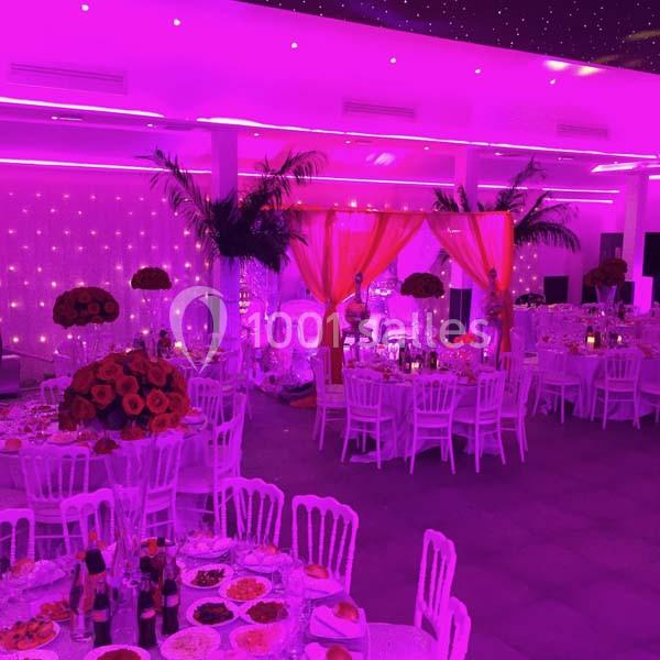 Salle de réception décorée avec des tables dressées, éclairage rose vif et rideaux ornés de fleurs rouges.