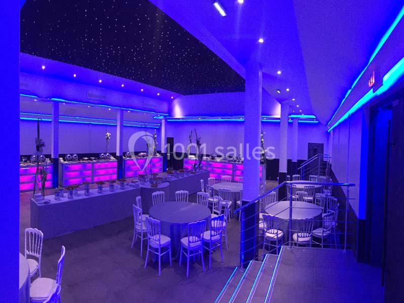 Salle de réception moderne avec éclairage LED bleu, tables rondes dressées et buffet en arrière-plan.