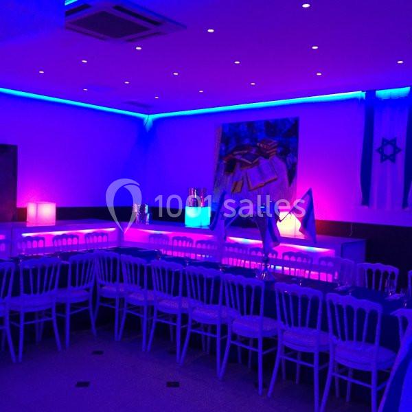 Salle décorée avec des lumières bleues et violettes, tables alignées avec chaises blanches, drapeaux visibles en arrière…