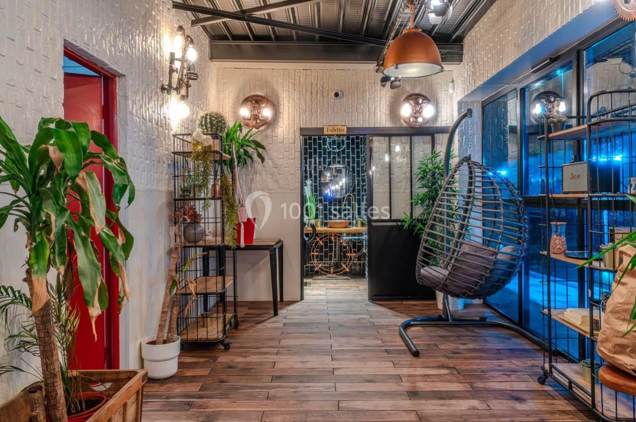 Couloir décoré de plantes, meubles en bois et luminaires modernes, menant à une pièce avec une table et des chaises.