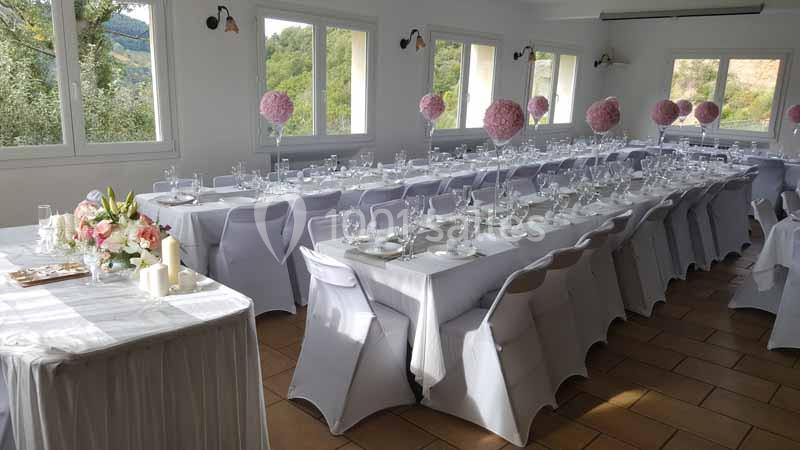 Salle de réception décorée avec des tables alignées, nappes blanches, verres, couverts et centres de table fleuris.