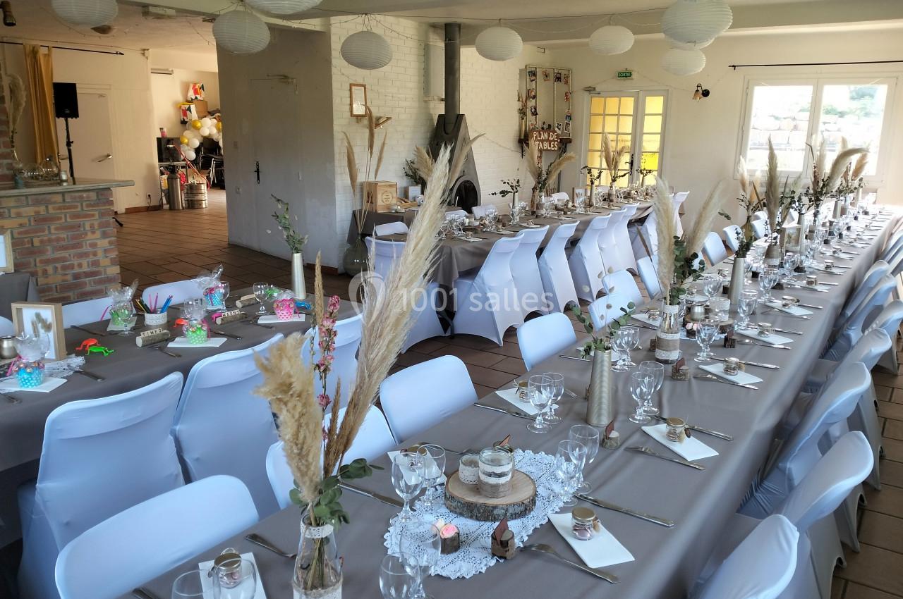 Salle décorée pour un événement avec longues tables dressées, nappes grises, chaises blanches et ornements naturels.