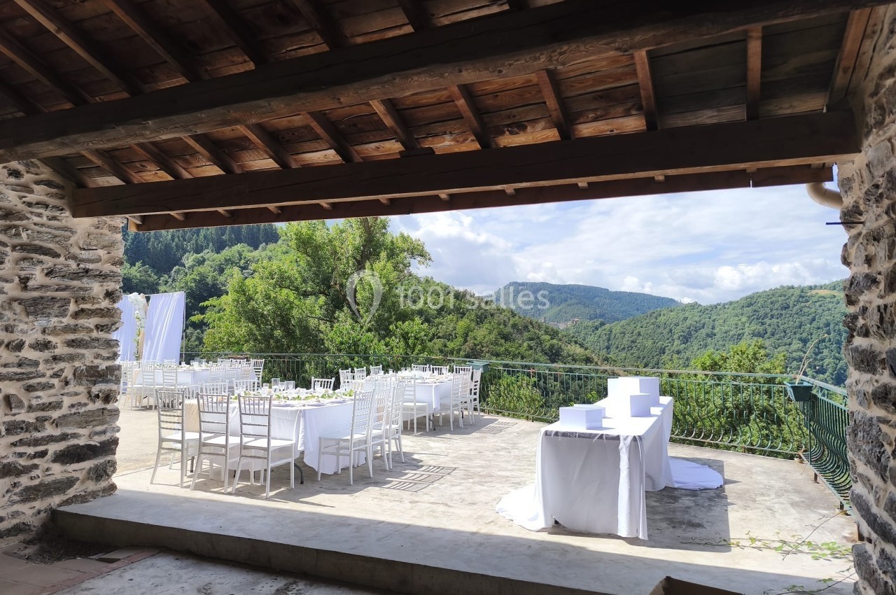 Terrasse en pierre avec tables et chaises blanches, offrant une vue sur des collines verdoyantes sous un ciel partiellement…