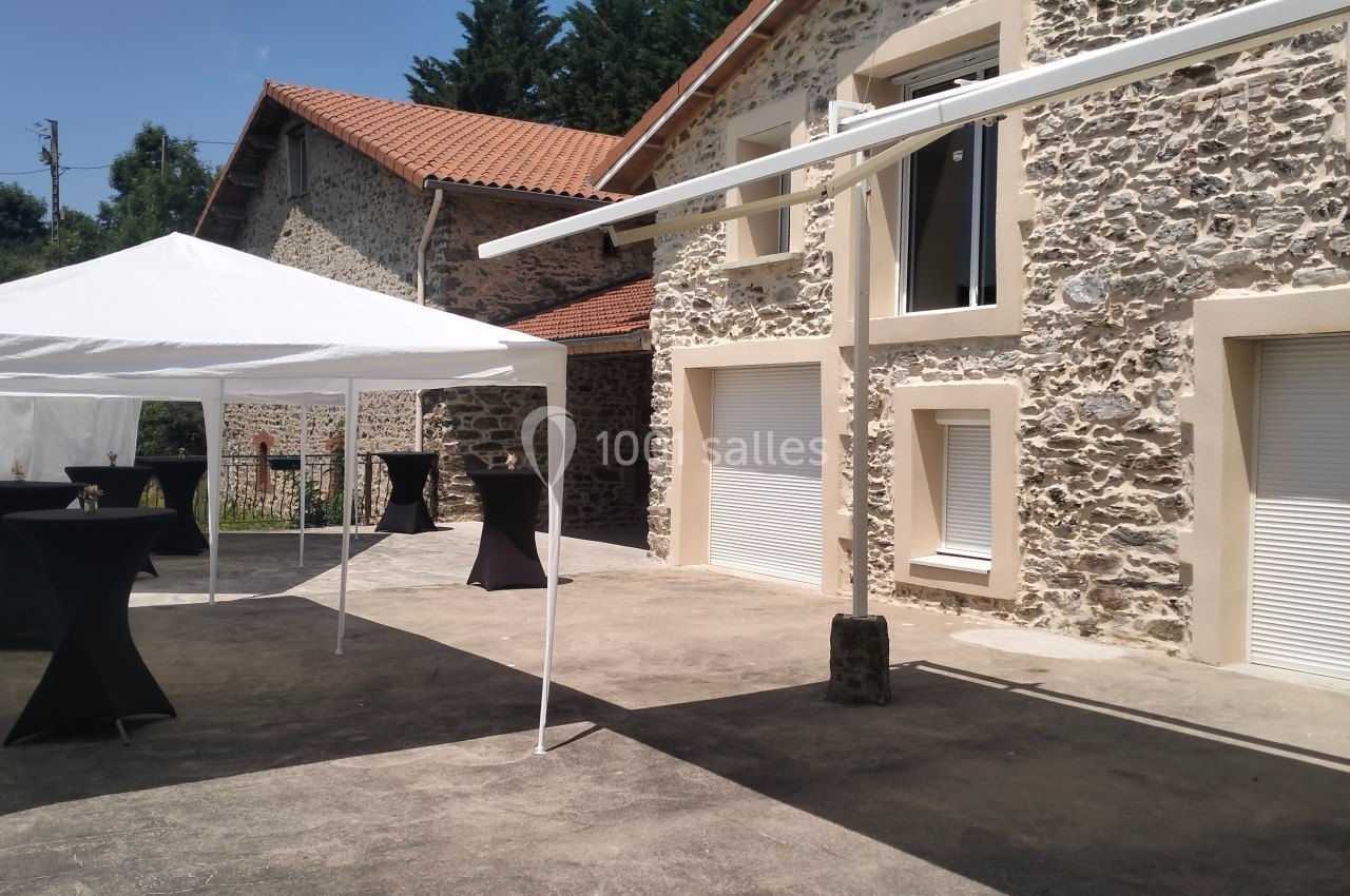 Cour extérieure avec des bâtiments en pierre, un barnum blanc et des tables hautes noires sur une terrasse ensoleillée.