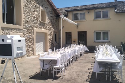Tables dressées pour un repas en extérieur devant une maison en pierre et crépi, avec une borne photo à gauche.
