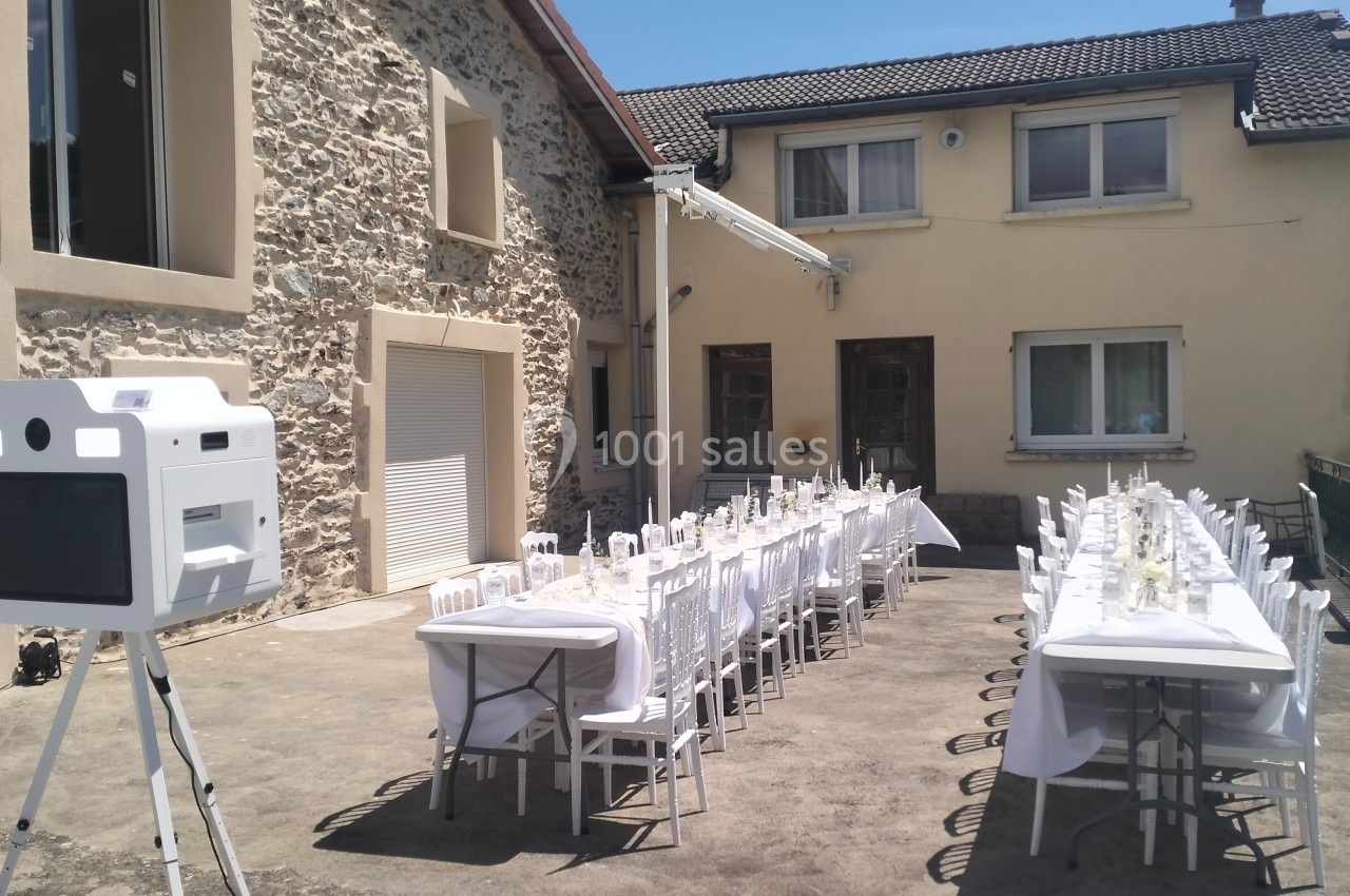 Tables dressées pour un repas en extérieur devant une maison en pierre et crépi, avec une borne photo à gauche.