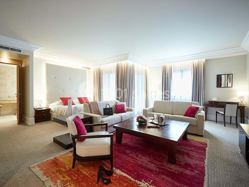 Chambre spacieuse avec lit double, coin salon, tapis coloré, grandes fenêtres et salle de bain attenante.