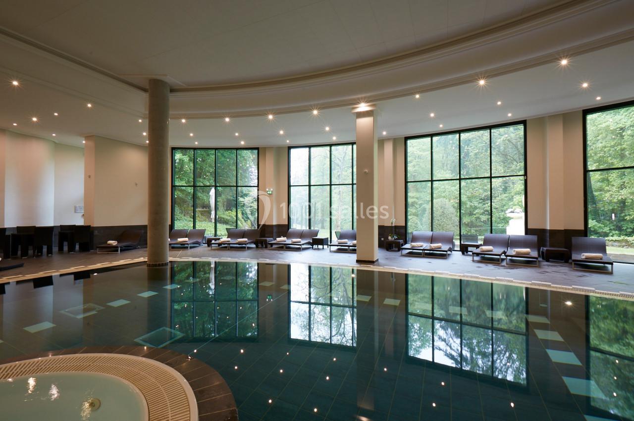 Piscine intérieure avec transats alignés près de grandes baies vitrées donnant sur un paysage verdoyant.