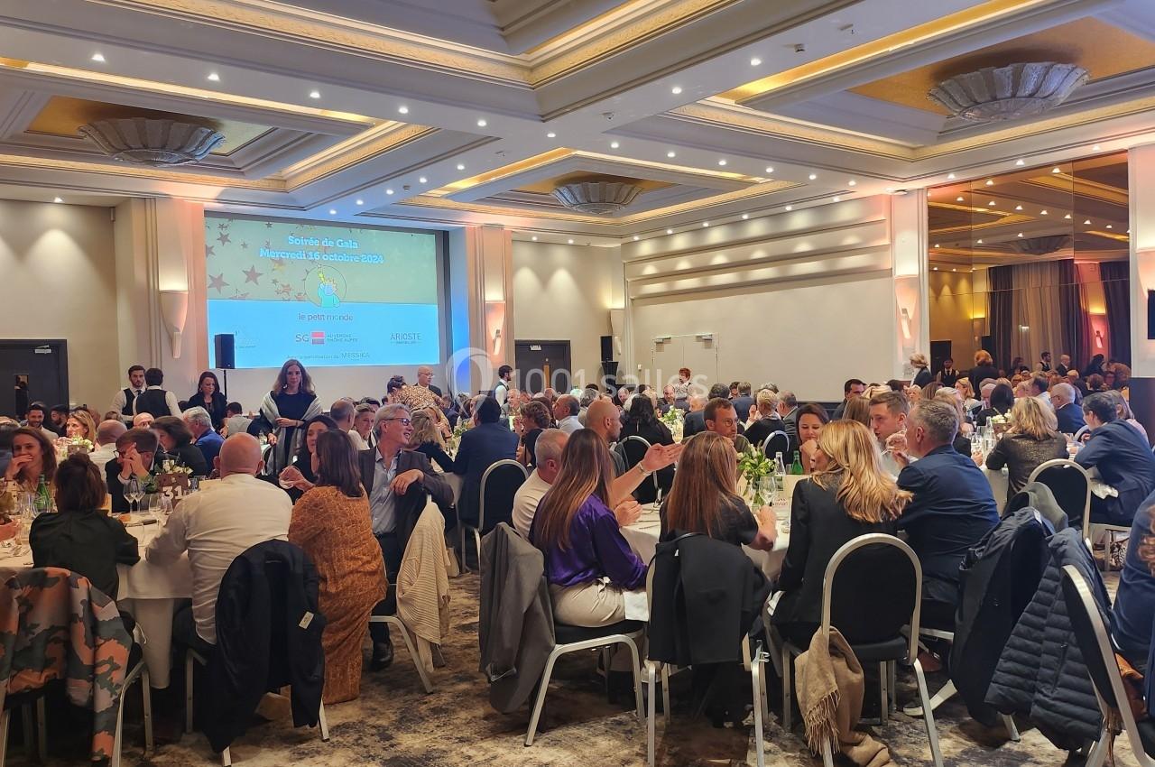 Salle de réception remplie de convives assis autour de tables, avec un écran projetant une présentation en arrière-plan.