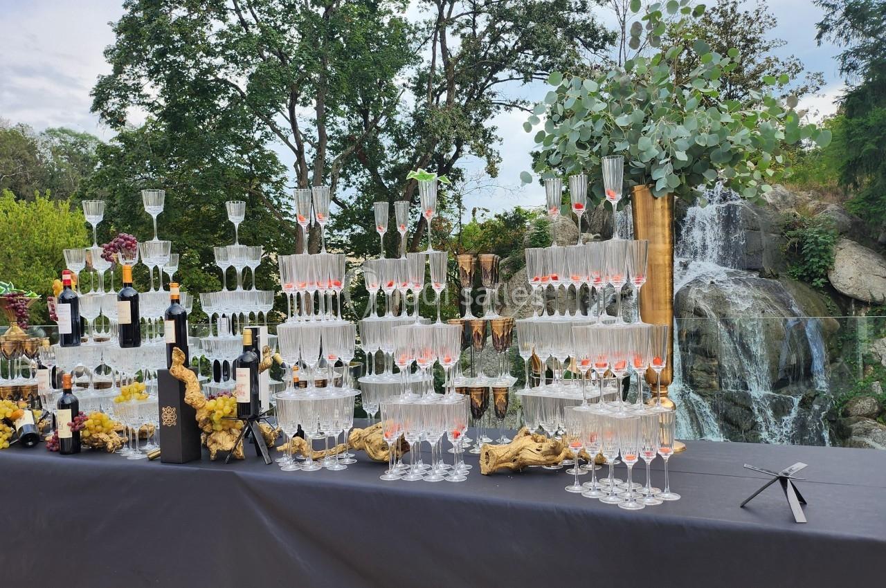 Table ornée de verres empilés, bouteilles de vin et décorations, avec une cascade et des arbres en arrière-plan.
