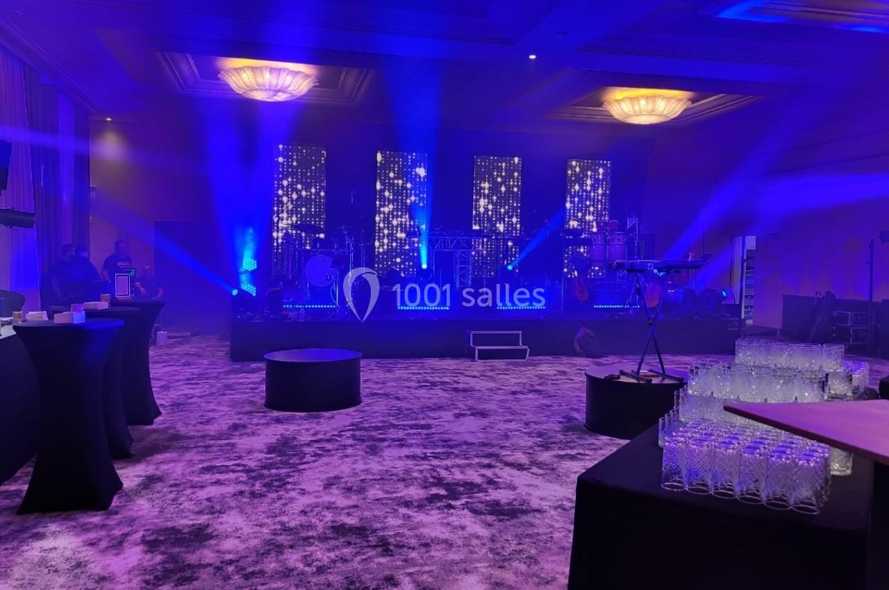 Salle de réception illuminée par des projecteurs bleus, avec une scène équipée d'instruments et des tables dressées.