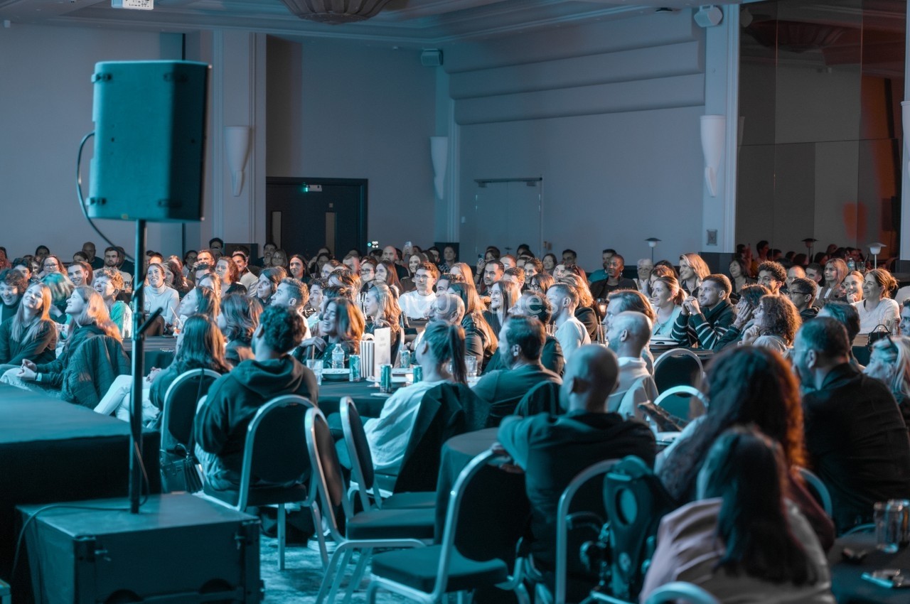 Un public assis dans une salle de conférence, attentif à une présentation sur une scène éclairée.