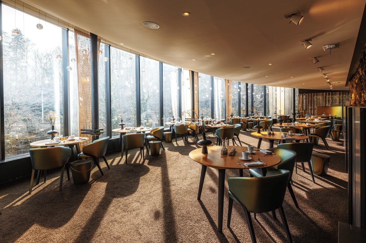 Salle de restaurant lumineuse avec de grandes baies vitrées, tables en bois et chaises modernes, vue sur un paysage boisé.