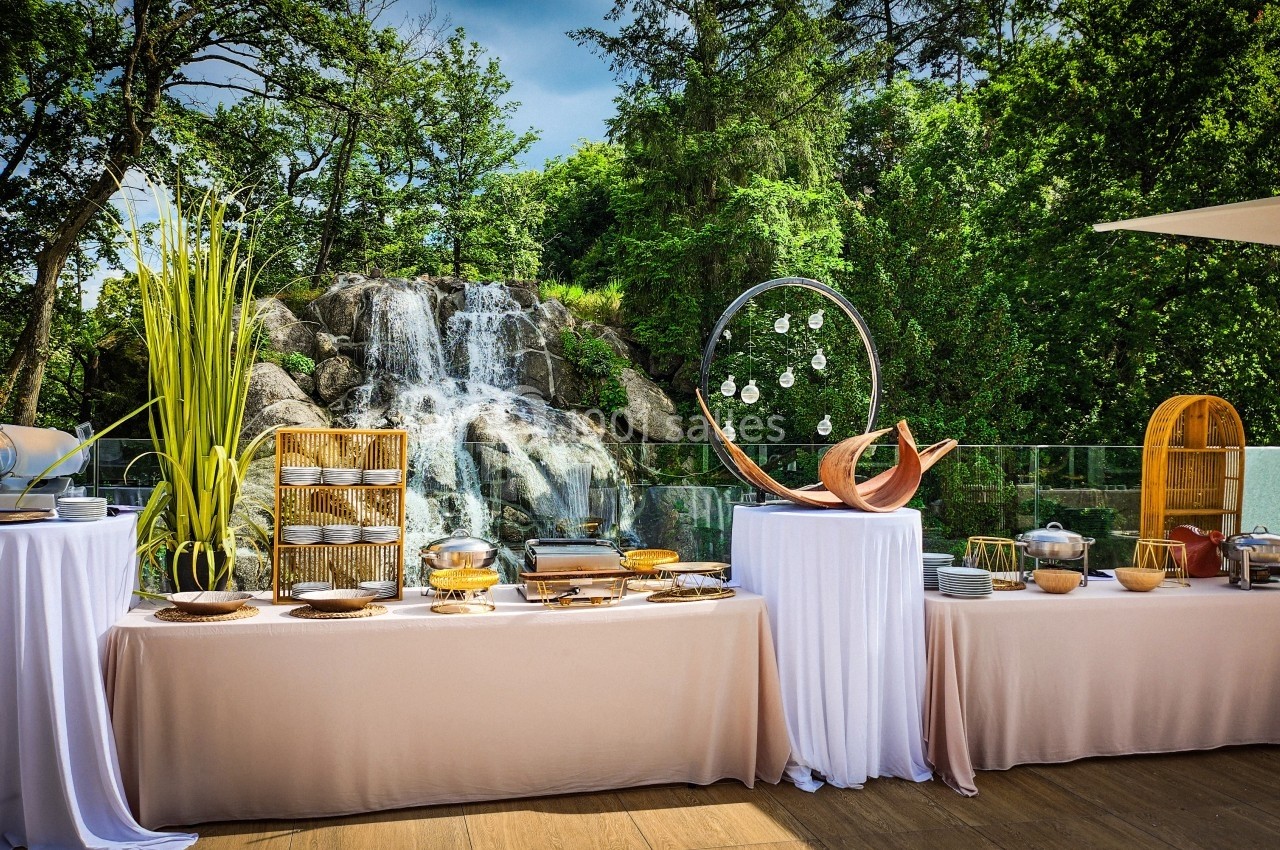 Buffet en plein air avec nappes blanches, plats variés et une cascade entourée de verdure en arrière-plan.