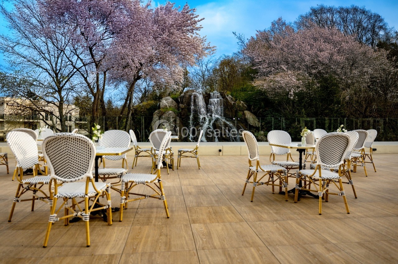 Terrasse avec chaises en rotin, tables dressées, arbres en fleurs et une cascade en arrière-plan.