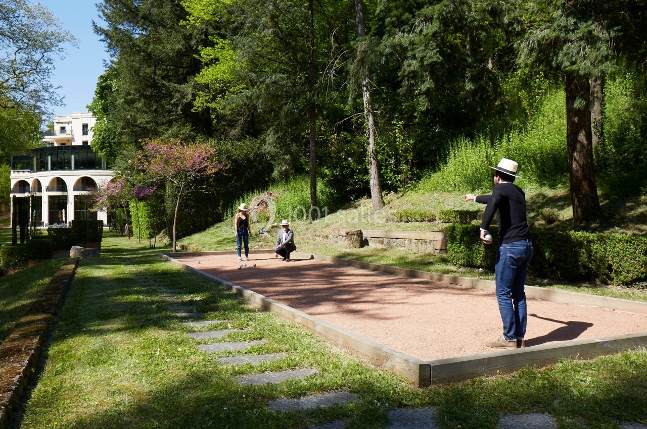 Personnes jouant à la pétanque sur un terrain ensoleillé entouré de verdure et d'arbres.