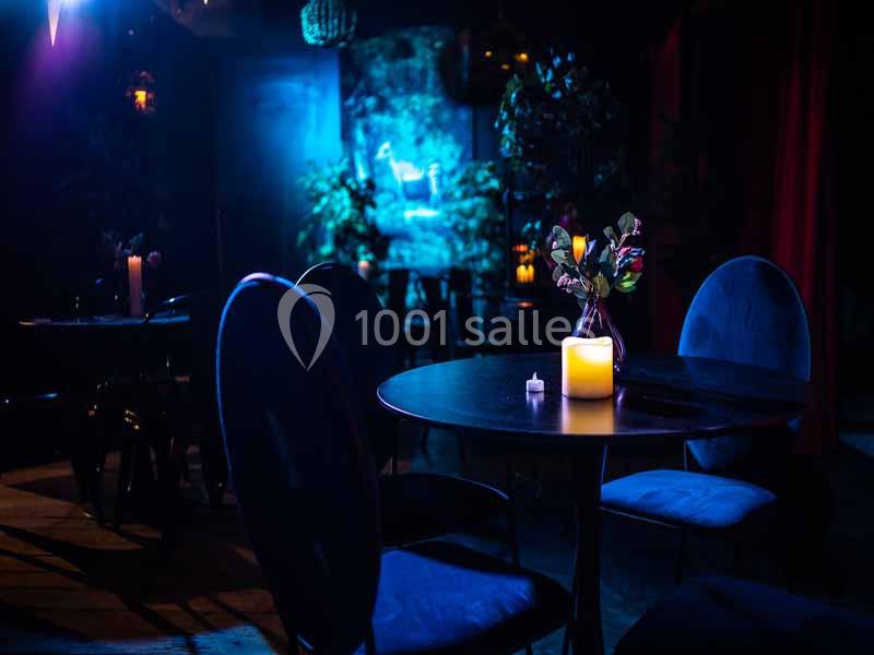 Salle sombre avec des tables rondes, des chaises bleues, une bougie allumée et une lumière bleutée en arrière-plan.